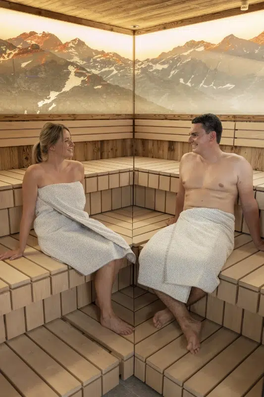 Zwei Personen entspannen sich in einer Sauna des Wellnesshotels Kaunertal und lächeln mit einem Bergwandbild im Hintergrund.