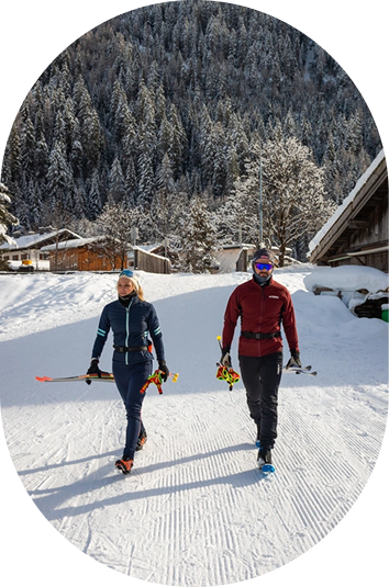 Zwei Personen gehen auf dem verschneiten Kaunertaler Winterboden mit ihren Skiern an Bäumen und Hütten vorbei.