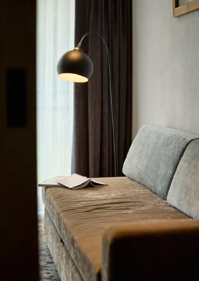 Eine Kaunertaler Familienzimmer-Stehlampe leuchtet auf ein aufgeschlagenes Buch auf einem grauen Sofa neben einem Vorhang.