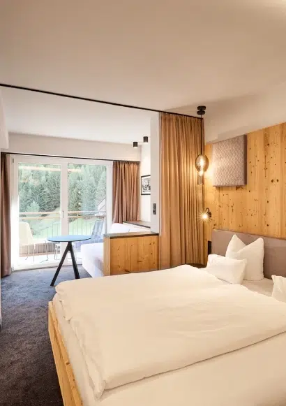 Helles Kaunertal Doppelzimmer mit Doppelbett, Sofa, Balkon und großem Fenster mit Blick auf Bäume.