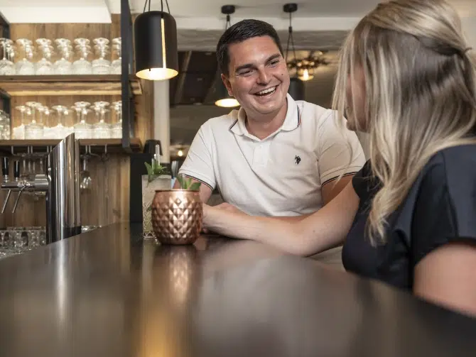Mann und Frau lächeln an der Bar eines Hotels im Kaunertal und plaudern über Getränke.