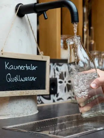 Eine Hand füllt eine Glasflasche mit Wasser aus einem Wasserhahn neben einem Kaunertaler Quellwasser Schild.