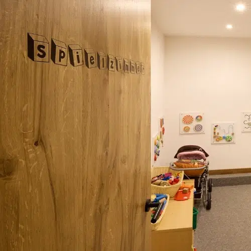 Die teilweise geöffnete Tür mit der Aufschrift Spielzimmer offenbart ein Hotel Kaunertal Spielzimmer mit Spielzeug und Kinderwagen.