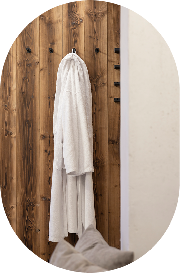 Weißer Bademantel, der an einer Holzwand im Wellnesshotel Kaunertal hängt, in der Nähe der Couch und der strukturierten beigen Oberfläche.