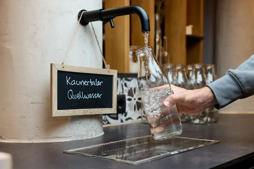 Eine Person füllt eine Glasflasche mit Wasser aus einem schwarzen Wasserhahn im Hotel Kaunertal.