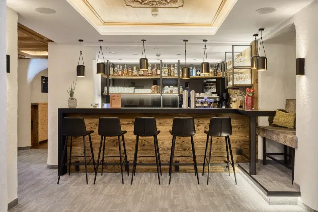 Moderne Bar im Hotel Kaunertal mit Holztresen, schwarzen Hockern und Flaschenregalen.