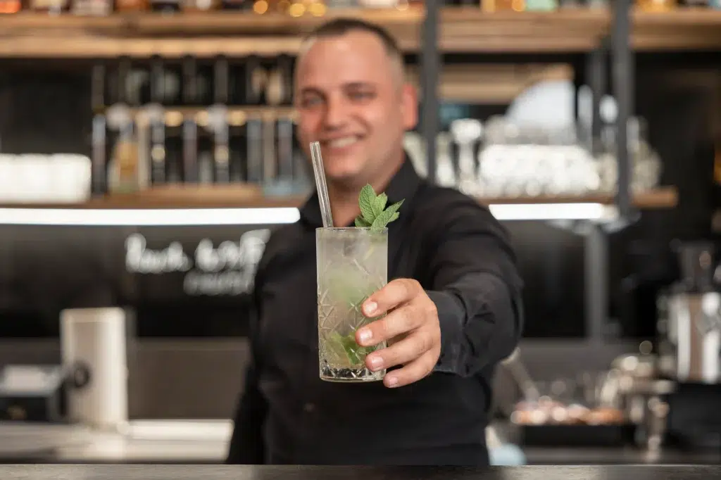 Der Barkeeper lächelt und hält einen kulinarisch inspirierten Cocktail mit Minzgarnitur in die Kamera.