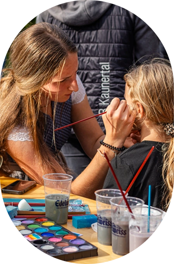 Eine Frau malt das Gesicht eines jungen Mädchens an einem Kunsttisch der Veranstaltungen Kaunertal im Freien.