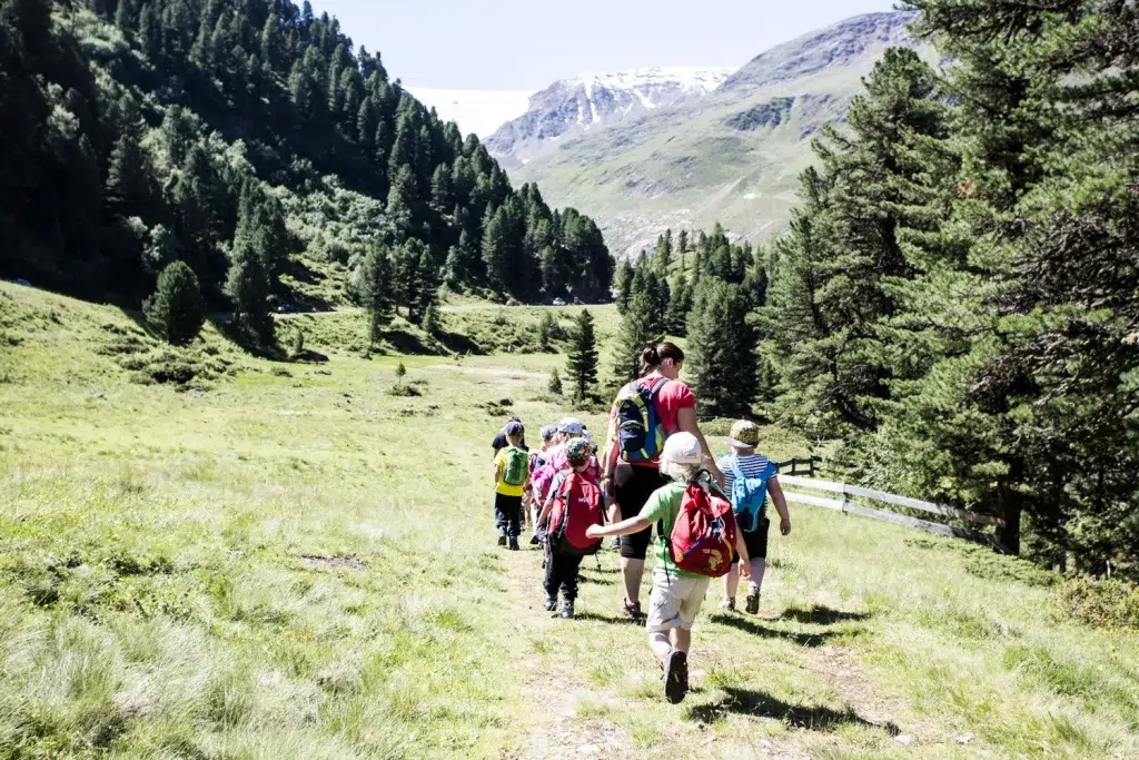 Eine Gruppe von Kindern wandert im Familienurlaub Kaunertal auf einem grasbewachsenen Bergweg, umgeben von Bäumen.