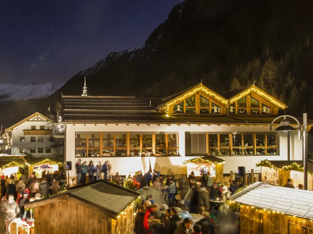 Ein festlicher Veranstaltungen Kaunertaler Markt bei Nacht mit Lichtern, Menschenmassen und verschneiten Bergen.
