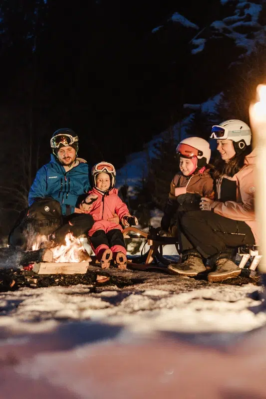 Eine Familie in Skikleidung genießt ein winterliches Kaunertaler Lagerfeuer in der verschneiten Nacht und lächelt gemeinsam.