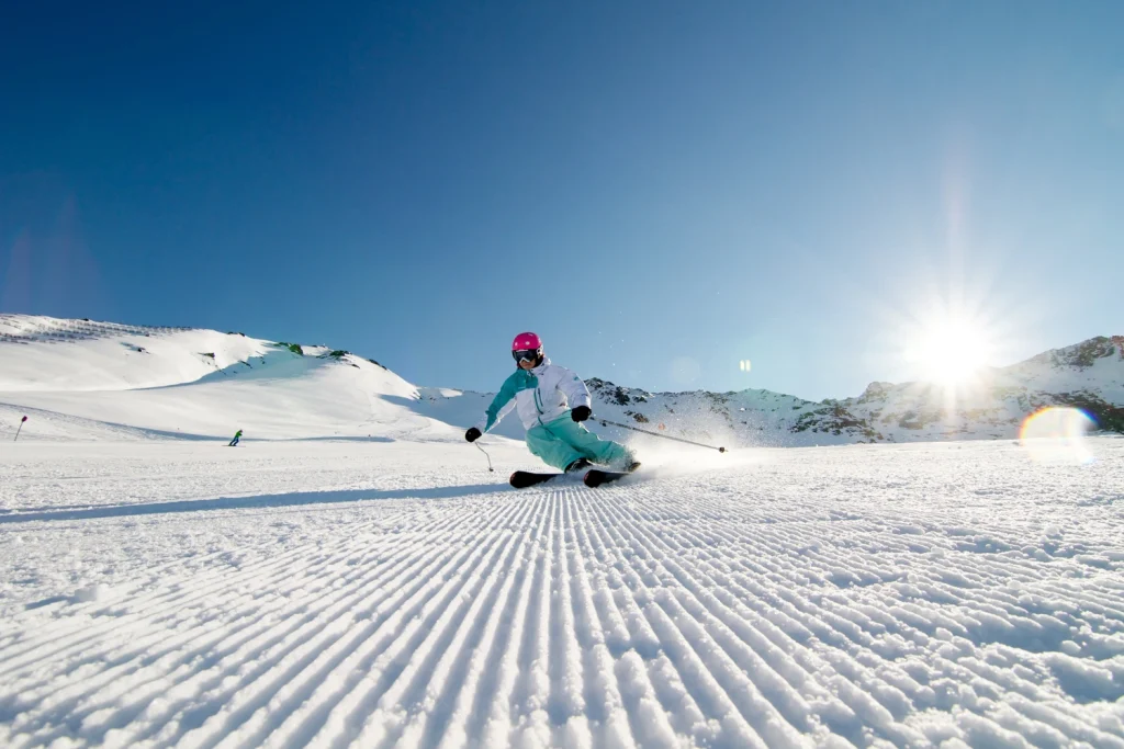 Ein Skifahrer in farbenfroher Ausrüstung carvt auf einer sonnigen, präparierten Winter-Kaunertaler Piste mit Bergen im Hintergrund.
