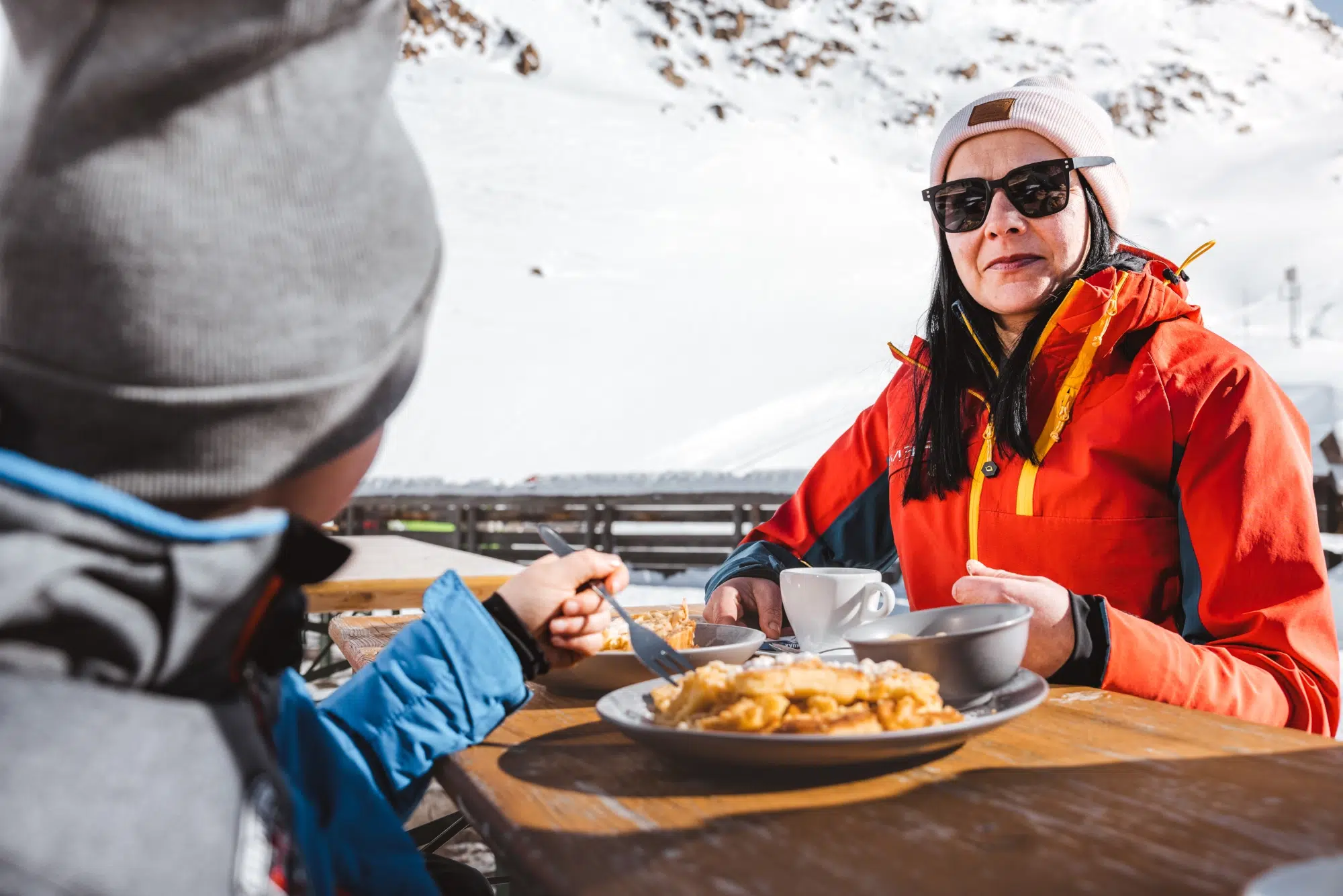 Frau mit Sonnenbrille und Kind speisen draußen in den verschneiten Bergen in der Nähe des Hotels Kaunertal.