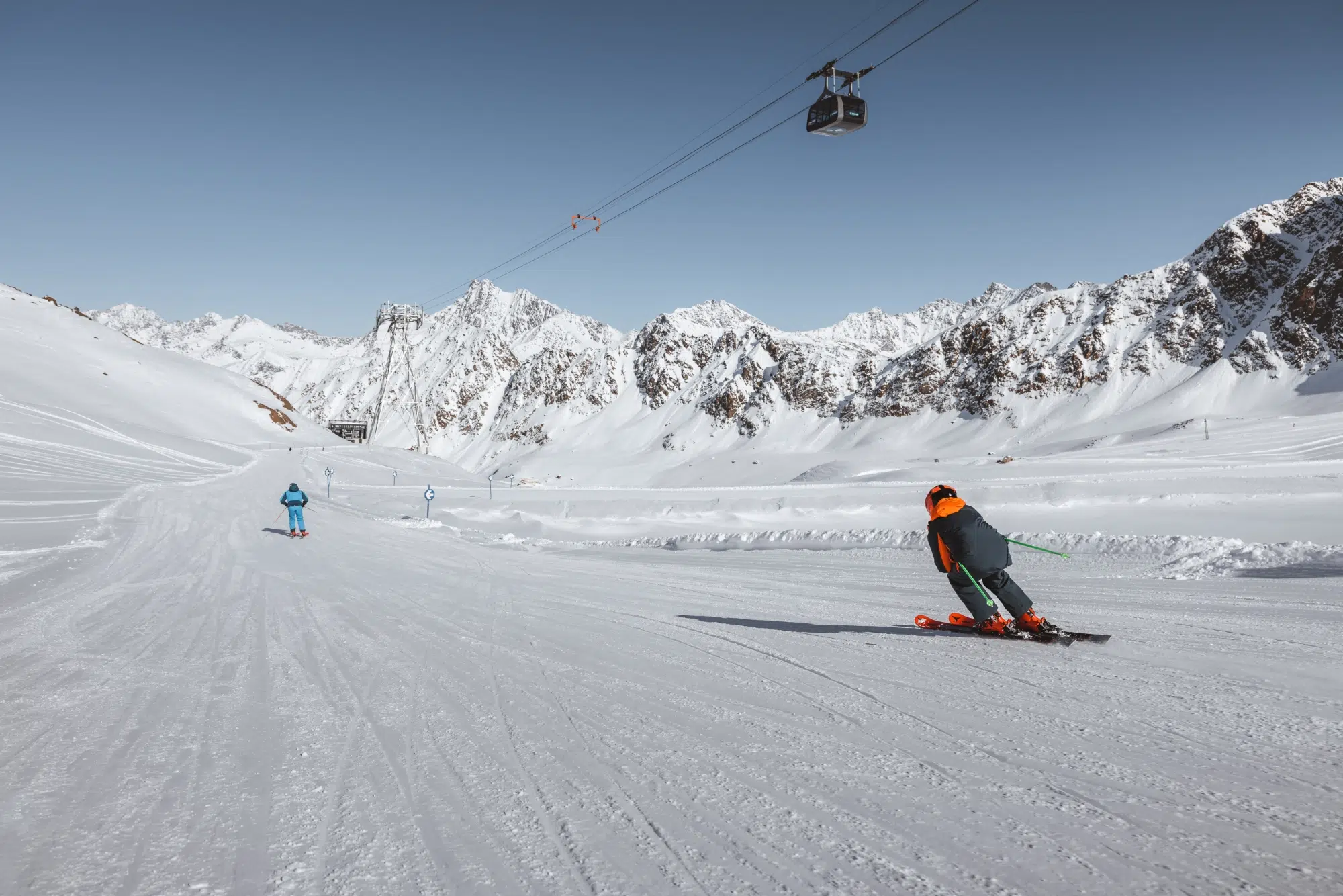Zwei Skifahrer auf einer verschneiten Piste in der Nähe des Hotels Kaunertal, mit Bergen und einer Seilbahn im Hintergrund.