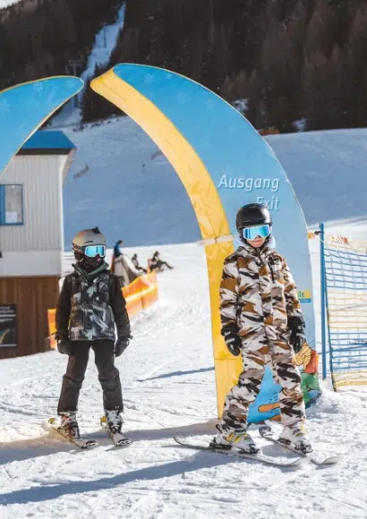 Zwei Kinder in Skiausrüstung stehen neben einem Kaunertaler Skipass-Ausgangsschild auf einer verschneiten Piste.