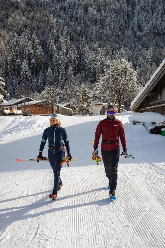 Zwei Skiträger gehen im Winter im Kaunertal auf Schnee, dahinter verschneite Hütten und Bäume.