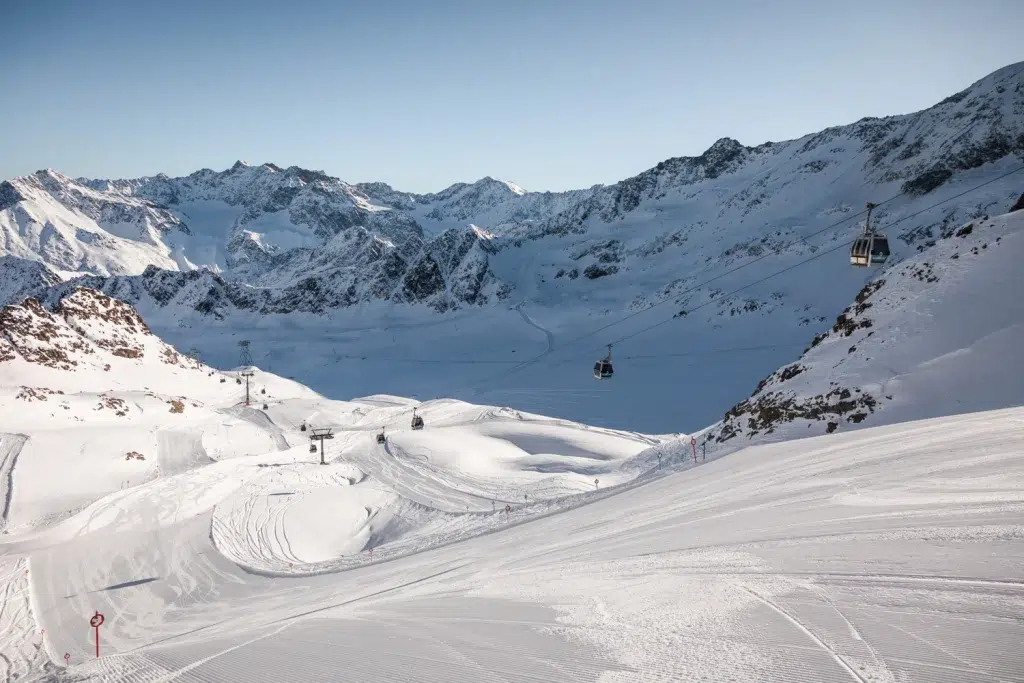 Eine verschneite Winter-Kaunertaler Skipiste mit Seilbahnen und fernen Gipfeln unter einem klaren Himmel.