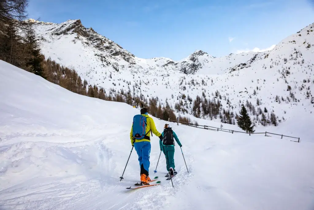 Zwei Personen fahren im Winter Kaunertal auf einer verschneiten Bergstrecke zwischen Bäumen und hohen Gipfeln bergauf.