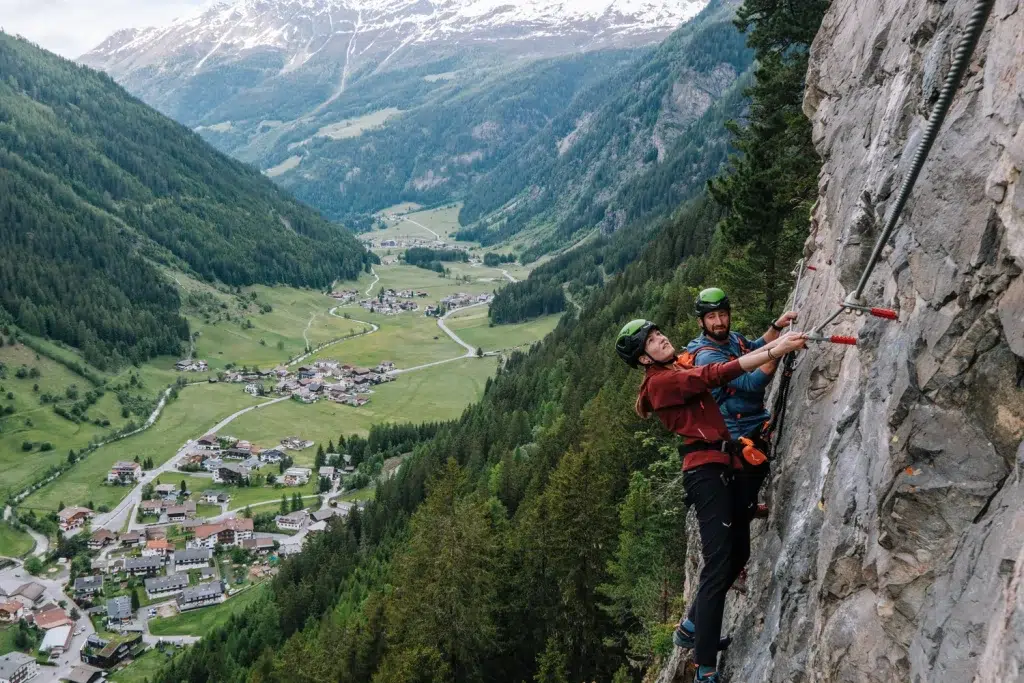 Zwei Bergsteiger erklimmen im Kaunertaler Sommer einen Berghang über einem malerischen Tal und fernen Gipfeln.