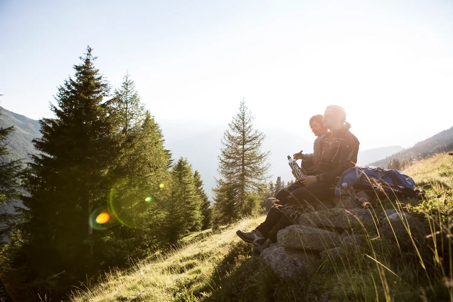 Zwei Menschen sitzen in einem sonnigen Kaunertaler Sommerwald auf einem Felsen, umgeben von Bäumen und Gras.